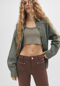 Cămașă cu carouri verzi și albe, top scurt din bumbac gri texturat, pantaloni strâmți maro cu capse argintii la buzunare. Păr lung și ondulat, fundal neutru.