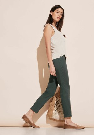 Haut sans manches gris clair, pantalon vert fuselé, chaussures brunes à enfiler, portant un sac en osier. Tenue simple et décontractée avec une coupe ample.