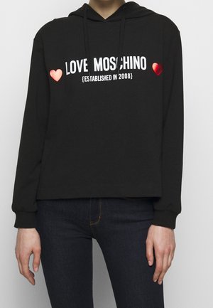Zwarte hoodie met voorop de tekst "LOVE MOSCHINO (OPGERICHT IN 2008)" omringd door twee rode hartjes, gedragen met donkere jeans.