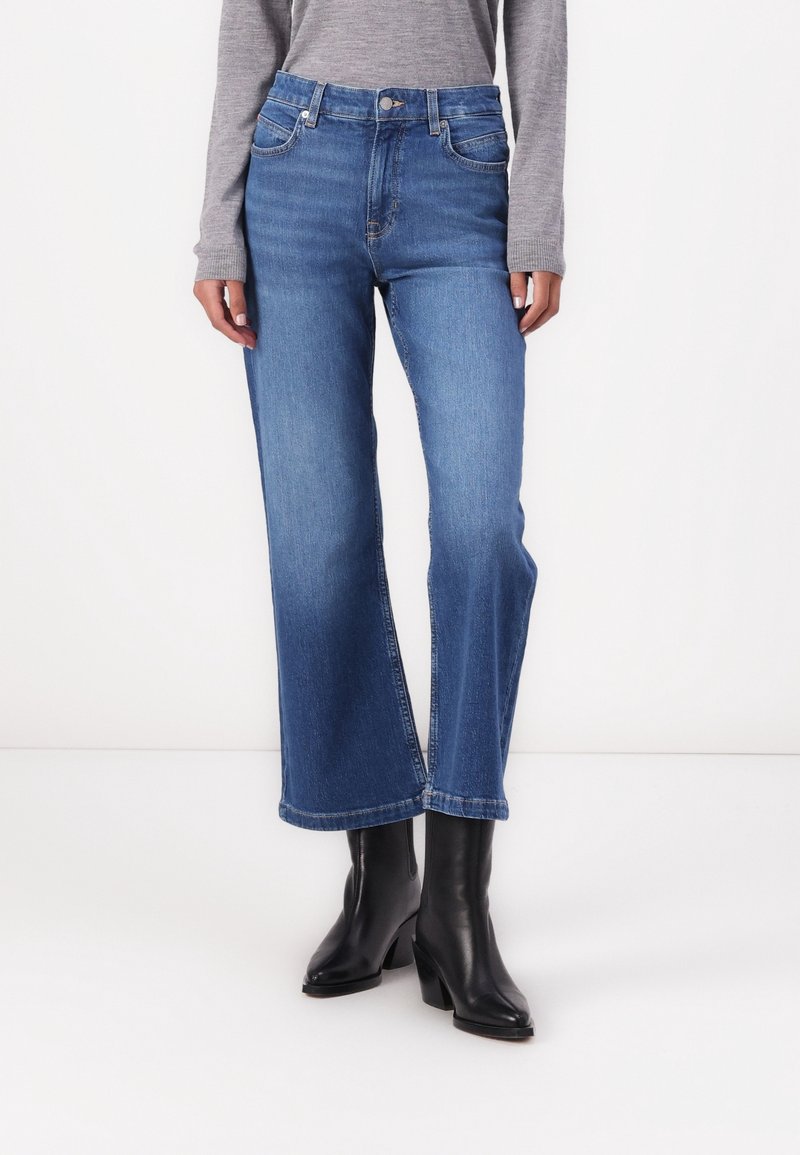Femme portant un jean droit en denim bleu taille mi-haute, un haut gris à manches longues et des bottines noires à talons, debout sur un fond blanc.