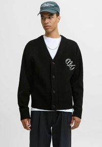 Schwarze übergroße Strickjacke aus strukturiertem Stoff, mit V-Ausschnitt, Knopfverschluss und gesticktem Logo auf der linken Seite.