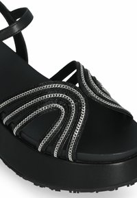 Sandale à plateforme noire avec des accents en chaîne argentée, des sangles courbées et une surface lisse texturée. La semelle est texturée pour une meilleure adhérence et stabilité.