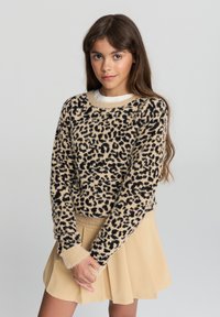 Ragazza con lunghi capelli castani che indossa un maglione leopardato beige e nero e una gonna beige, in piedi davanti a uno sfondo chiaro.