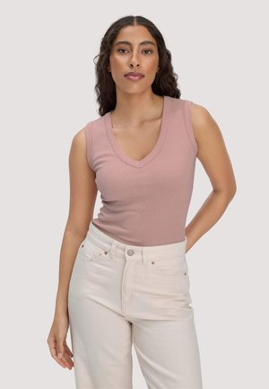 Frau mit langen lockigen Haaren, trägt ein staubrosa ärmelloses V-Ausschnitt-Oberteil und hoch taillierte cremeweiße Hose, posiert vor einem schlichten Hintergrund.