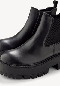 Bottines en cuir noir avec semelles épaisses à crampons, panneaux latéraux élastiques et languettes sur fond blanc.