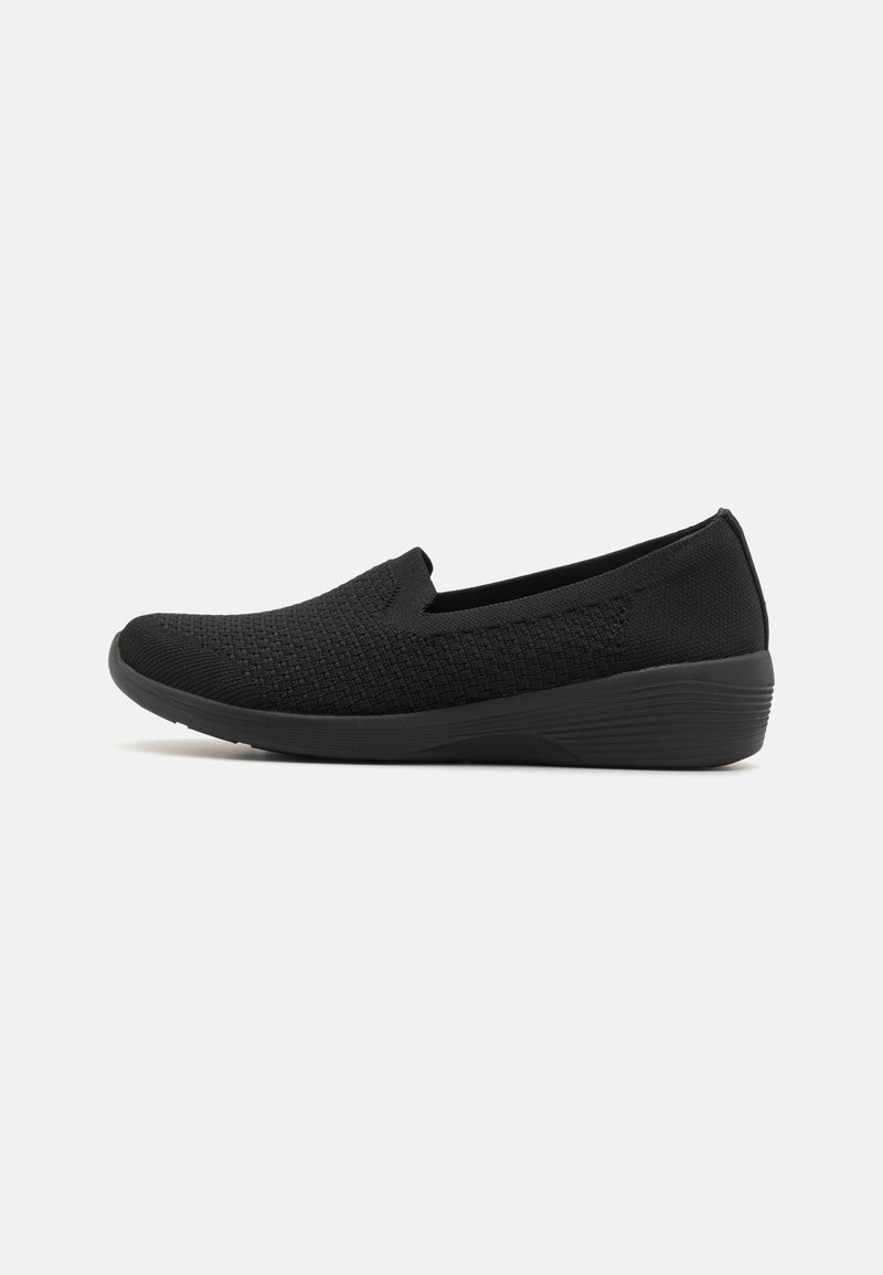 Chaussure noire sans lacets avec dessus en maille texturée et semelle compensée basse, conçue pour le confort et un usage décontracté.
