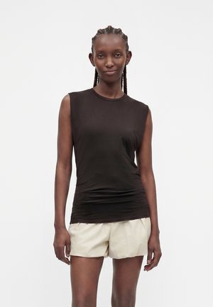 Femme aux cheveux tressés portant un haut noir sans manches et un short beige clair, debout devant un fond clair uni.