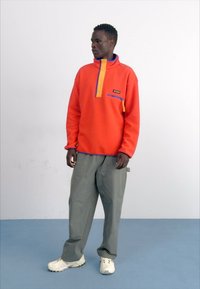 Rød fleece-pullover med høj krave, blå detaljer og en lynlås-lomme, parret med løstsiddende grå bukser og hvide sneakers.