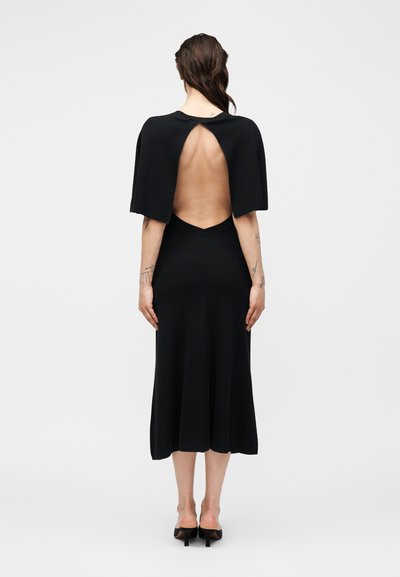 Γυναίκα με μακριά καστανά μαλλιά που φοράει μαύρο μίντι φόρεμα με γυμνή πλάτη και μανίκια σαν κάπα, στέκεται μπροστά σε ένα απλό φωτεινό φόντο.