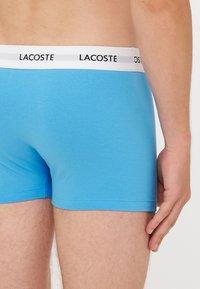 Boxer shorts en coton bleu avec une taille blanche portant le logo "Lacoste" en noir, texture lisse et silhouette ajustée.