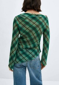 Persona con capelli scuri lunghi fino alle spalle, che indossa una maglietta a maniche lunghe a quadri verdi e jeans blu, vista di spalle su uno sfondo neutro.