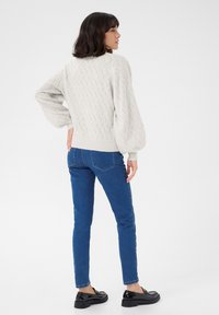 Pull en tricot texturé gris clair avec des manches bouffantes, associé à un jean bleu ajusté et des mocassins noirs, vu de dos sur un fond neutre.