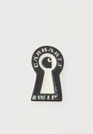 Black and white enamel pin shaped like a keyhole with "Carhartt" above and "WIP" below a central abstract eye logo.