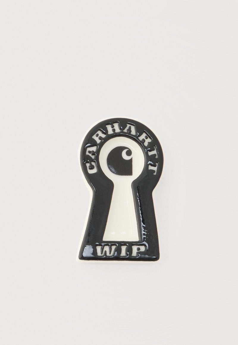 Καρφίτσα από σμάλτο σε ασπρόμαυρο χρώμα, σχήμα κλειδαρότρυπας με "Carhartt" πάνω και "WIP" κάτω από ένα κεντρικό αφηρημένο λογότυπο ματιού.