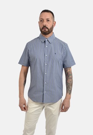 Uomo con barba e tatuaggi che indossa una camicia a righe verticali bianche e blu a maniche corte e pantaloni chiari, in piedi davanti a uno sfondo semplice.
