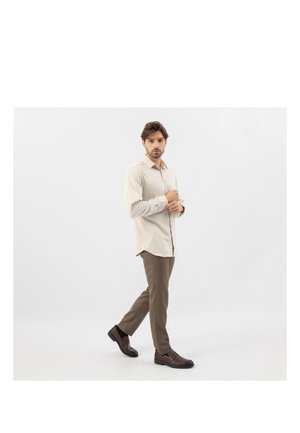 Uomo che cammina in avanti indossando una camicia beige, pantaloni marroni e scarpe eleganti marrone scuro su uno sfondo bianco semplice.