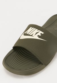 Maslinasto zelena klizna sandal koja je napravljena od teksturiranog sintetičkog materijala. Ima široku traku s bijelim Nike logom i rebrastom unutrašnjošću za prianjanje.