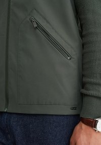 Temno zelena jakna z diagonalnim žepom na zadrgo, textured pleteni rokav, modri kavbojke in ročna ura z rjavim usnjenim paščkom.