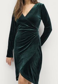Grünes Samtwickelkleid mit langen Ärmeln und V-Ausschnitt. Es verfügt über ein drapiertes Design und einen asymmetrischen Saum für eine figurbetonte Silhouette.
