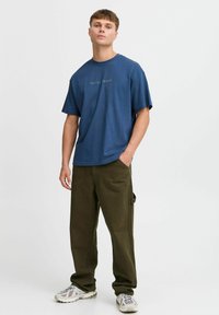 Camiseta de algodón azul con un diseño impreso que dice "JUEGA CONMIGO, MEMORIA", combinada con pantalones de corte holgado en verde oliva y zapatillas multicolores.
