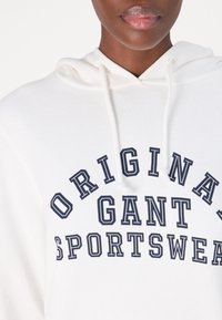 Sudadera blanca con texto impreso en navy "ORIGINAL GANT SPORTSWEAR." Cuenta con capucha ajustable y una tela texturizada.
