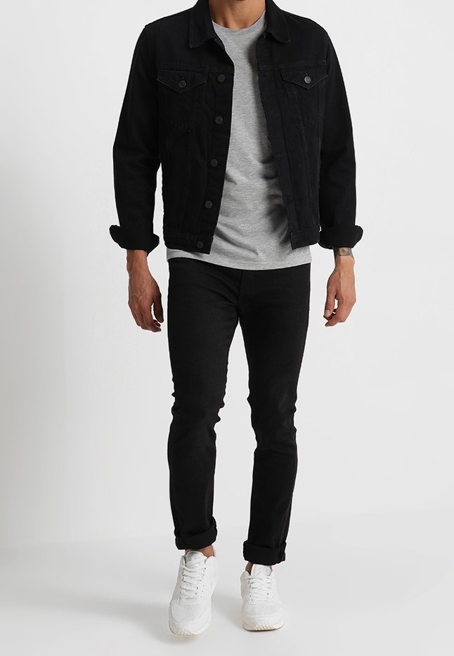Homme portant une veste en jean noire, un t-shirt gris, un pantalon noir ajusté et des baskets blanches sur un fond gris uni.