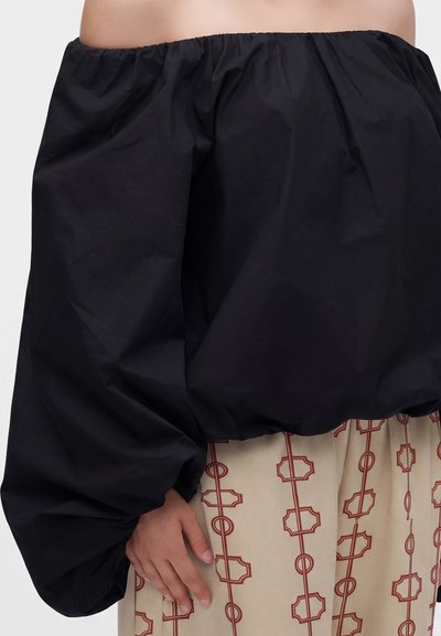 Μαύρο τοπ off-the-shoulder με φουσκωτά μανίκια συνδυασμένο με μπεζ παντελόνια με κόκκινα γεωμετρικά μοτίβα αλυσίδας.