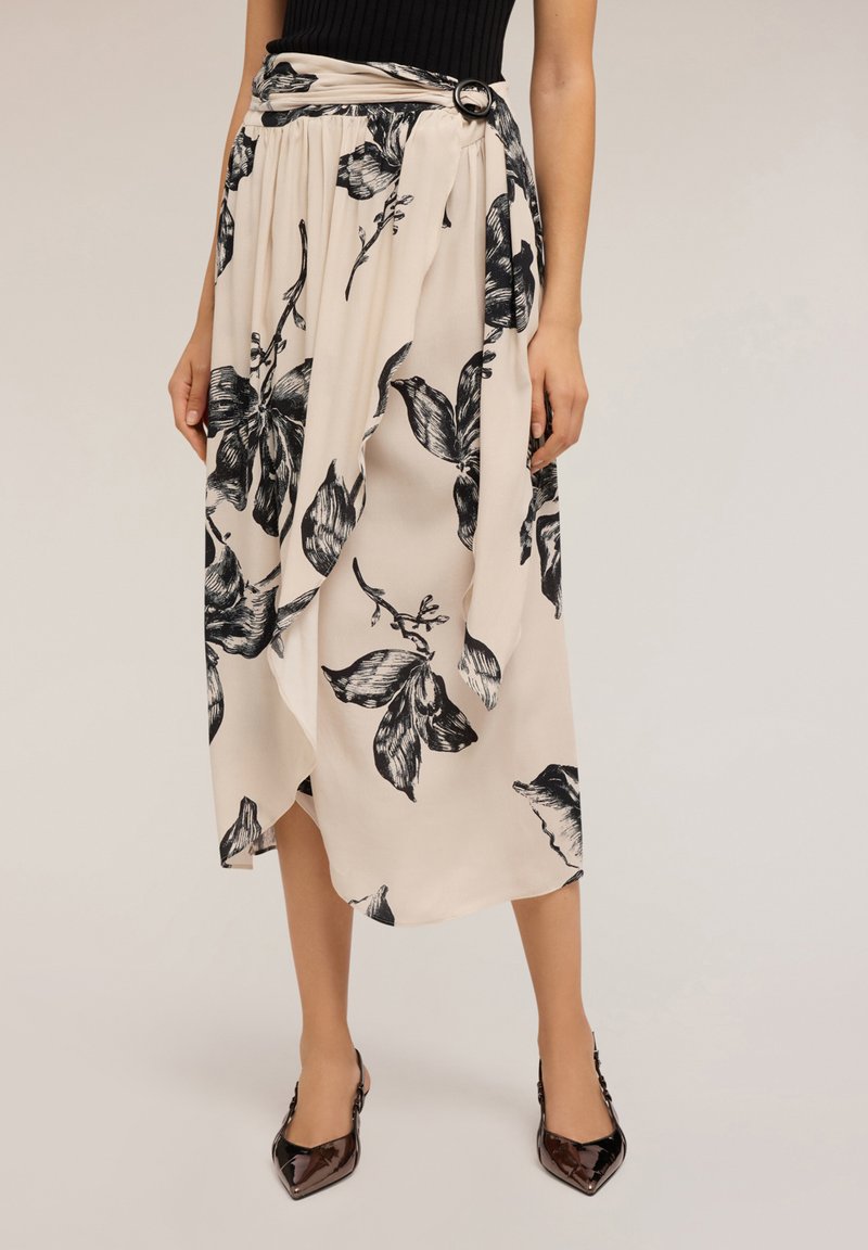 Gonna avvolgente beige con stampa floreale nera, vita arricciata con fibbia, indossata con un top nero e tacchi a punta lucidi su sfondo chiaro.