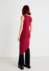 Vestido longo, sem mangas, na cor borgonha, com uma silhueta ajustada e bainha assimétrica, combinado com calças pretas de perna larga e sapatos pretos robustos.