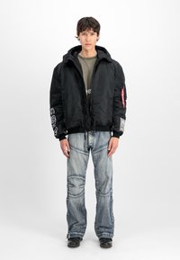 Melna izolēta bombers jakas ar kapuci, aprocēm un rāvējslēdzēju kabatām. Iezīmēta ar baltām un sarkanām akcentiem, kopā ar gaismas mazgājumiem džinsiem.