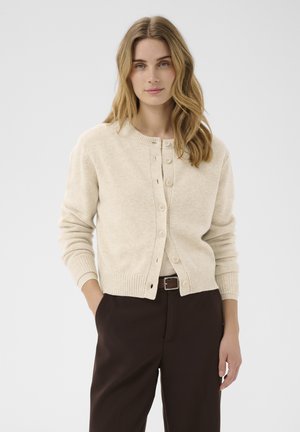 Jeune femme aux cheveux blonds ondulés portant un cardigan beige boutonné et un pantalon marron foncé, debout avec une main dans la poche.