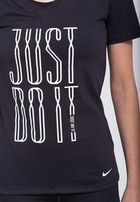 Camiseta de algodón negra con la frase "JUST DO IT" en letras blancas y en negrita. Mangas cortas, diseño de cuello en V, con un pequeño logo de Nike en el dobladillo.