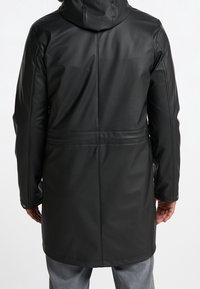 A person wears a long black coat with a hood, seen from the back. The coat is smooth and tailored, contrasted by light denim trousers.