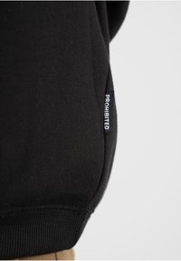 Sudadera negra con un ajuste ceñido, que presenta una pequeña etiqueta bordada que dice "PROHIBIDO" en el lateral. Textura de algodón suave.