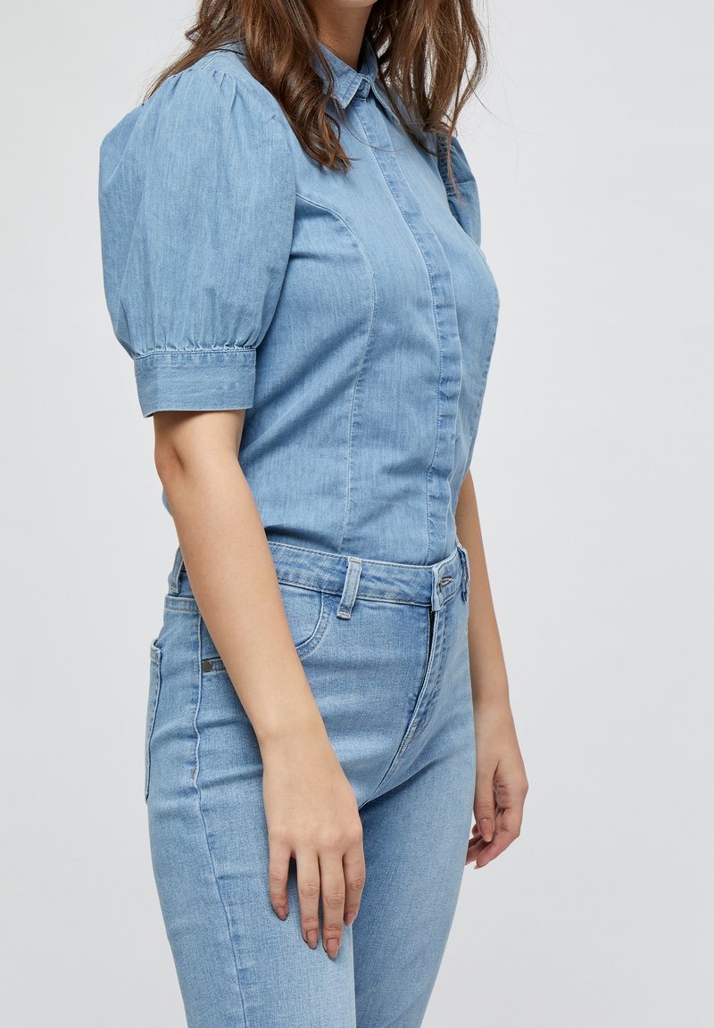Femme portant une chemise en denim bleu clair à manches bouffantes et un jean taille haute assorti, debout devant un fond uni.