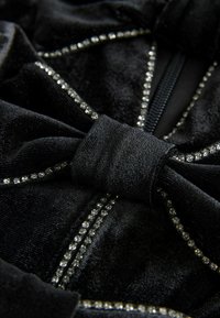 Mustan kankaan yksityiskohta, jossa on noita, ja jonka saumoissa on kiiltävät rhinestone-koristeet. Kankaalla on pehmeä rakenne ja hienovarainen kiilto.