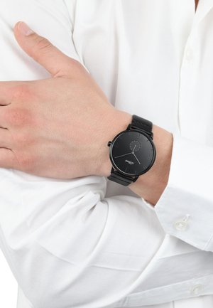 Schwarze analoge Uhr mit rundem Zifferblatt und minimalistischer Gestaltung, ausgestattet mit einem Lederarmband und silbernen Akzenten, getragen an einem nackten Handgelenk.