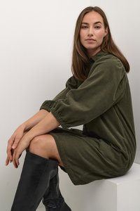 Olíva zöld kordbársony ruha, voluminous ujjakkal és laza fazonnal, térd fölé érő fekete bőr csizmákkal párosítva. Egyszerű és texturált kialakítás.