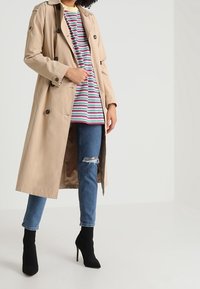 Trench beige avec des boutons croisés, superposé sur une chemise rayée, associé à un jean bleu déchiré et des bottes à talons noirs.