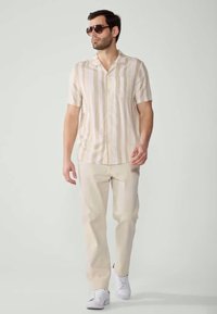 Hellbeige gestreiftes kurzärmliges Hemd kombiniert mit hellbeigen Hosen und weißen Sneakers, getragen mit dunklen Sonnenbrillen. Glatte Stofftextur.