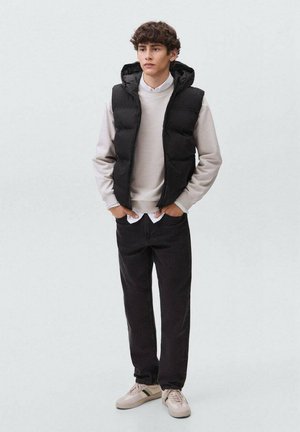 Gilet puffy nero sopra una felpa grigia chiara, jeans scuri e sneakers beige. Presenta un cappuccio e tasche laterali, con una vestibilità comoda.