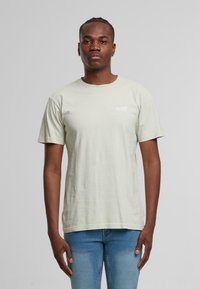 Hellgrünes Baumwoll-T-Shirt mit kurzen Ärmeln, Rundhalsausschnitt und einem kleinen weißen Logo auf der linken Brust. Lässige Passform, glatte Textur.