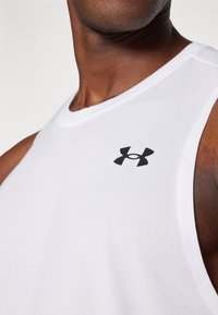 Débardeur de sport sans manches blanc en tissu lisse, avec un logo Under Armour noir sur le côté gauche.