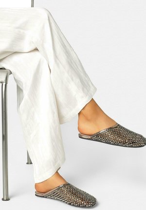 Persona con pantalones blancos sentada en una silla de metal, con los pies en zapatos planos metálicos de tejido entrelazado, un pie ligeramente levantado.