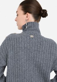 Maglione a collo alto a coste grigio, con un alto colletto e dettagli in metallo dorato sulle spalle, che mette in evidenza la texture e il design aderente.