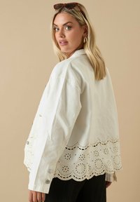 Vit bomullskavaj med en scalloped broderad fåll och knappstängning. Har långa ärmar och en avslappnad passform.