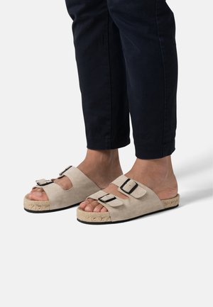 Sandales en daim beige avec deux brides réglables en cuir noir, semelle en corde tissée et dessous plat en caoutchouc noir, montrant des pieds nus.
