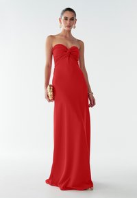 Strapless rode avondjurk met een gedraaide knoopdetail bij de bovenzijde, vloeiende, gladde stof en een aansluitende silhouet. Gouden clutchaccessoire.