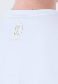 Camiseta de algodón blanca con cuello redondo. Presenta un pequeño logo en relieve en forma de 'K' en la parte posterior. Textura suave.