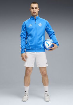 Jonge man staand, gekleed in een blauw jack met ritssluiting en witte korte broek, houdend een blauw-witte voetbal met het Puma-logo.